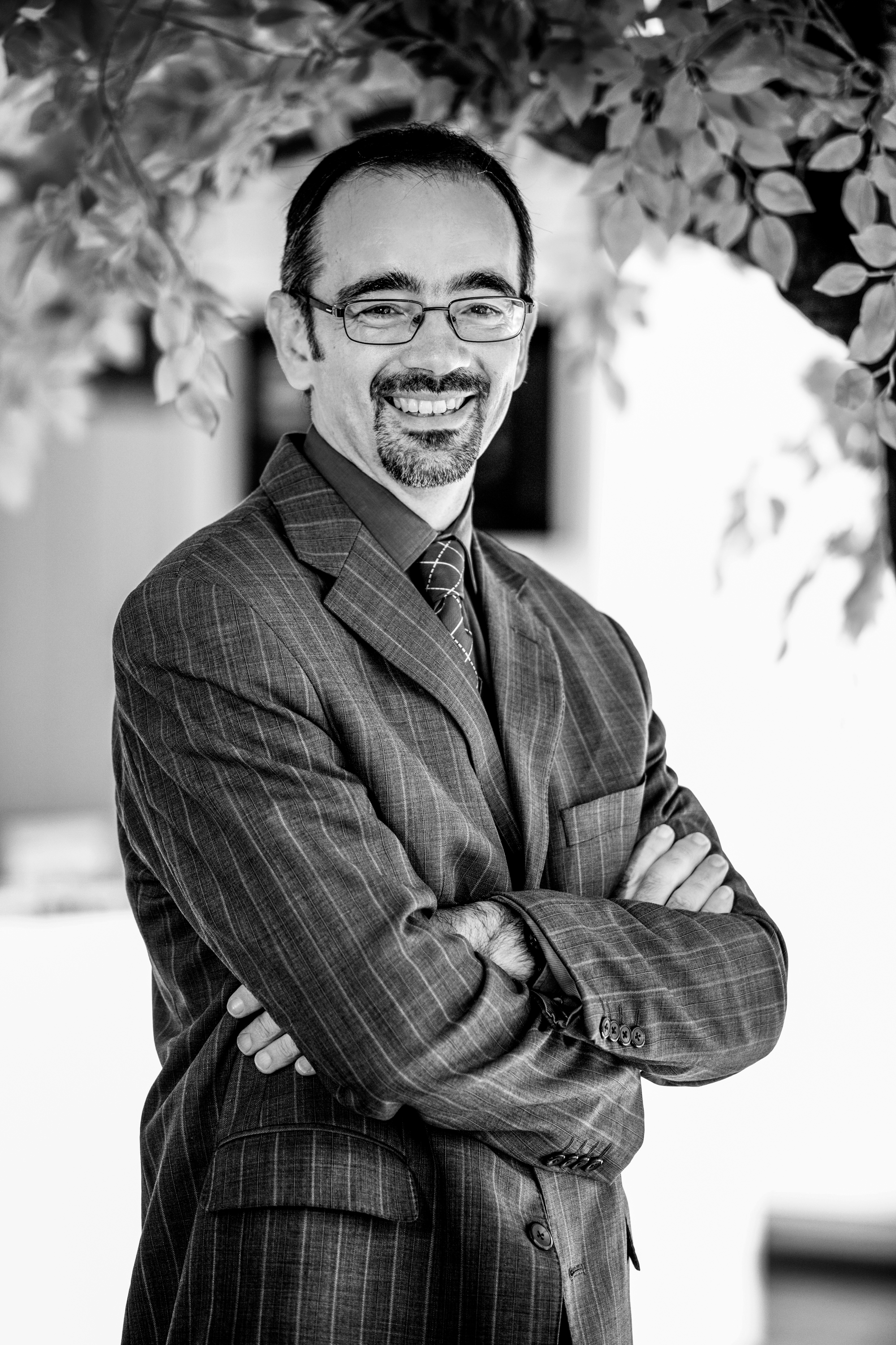 Black-and-white portrait of Christian Chevalier, smiling, arms crossed, beneath a canopy of leaves.